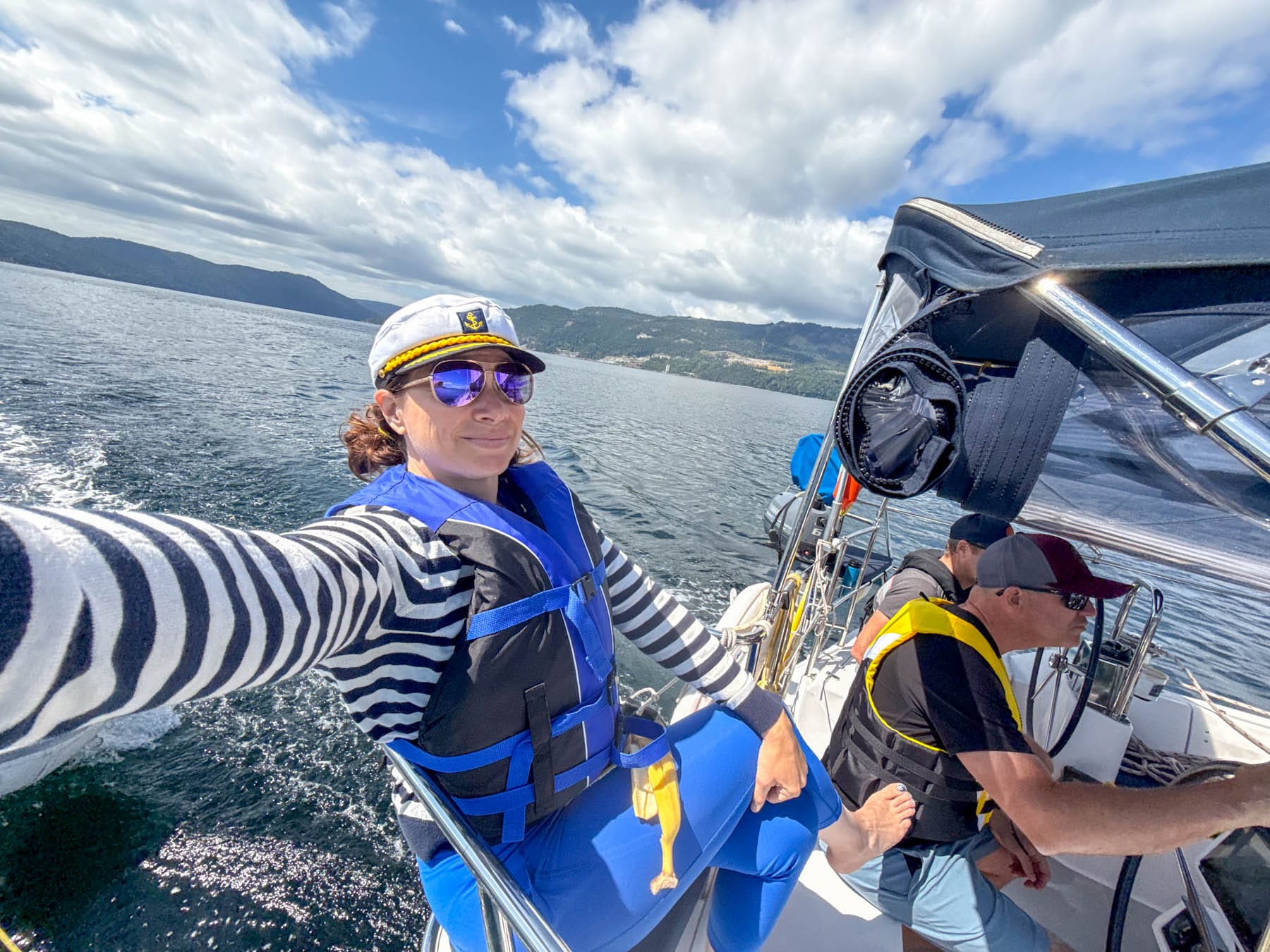 Jami Savage in captain's hat and sunglasses on a boat, wearing a blue life vest, with men and mountains in background