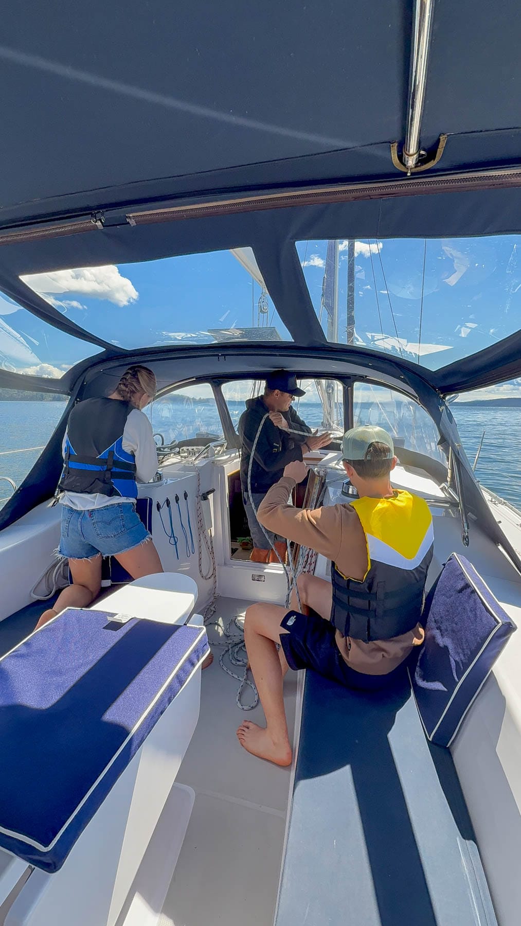 Three people in a sailboat cockpit, with two standing and one seated, all wearing life vests