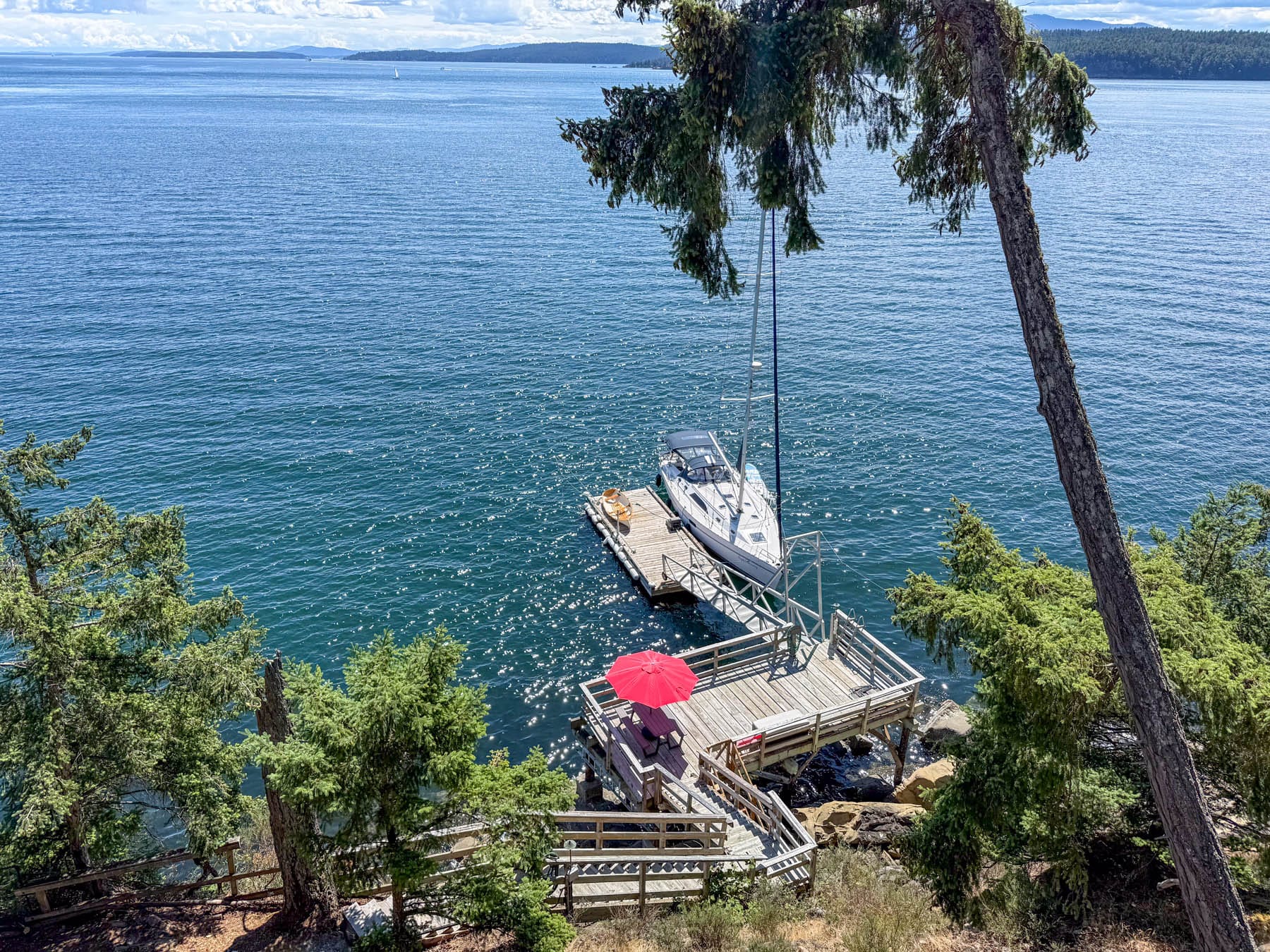 overhead view of a dock with a sailboat, kayak, red umbrella, and stairs leading to shimmering blue water with green trees