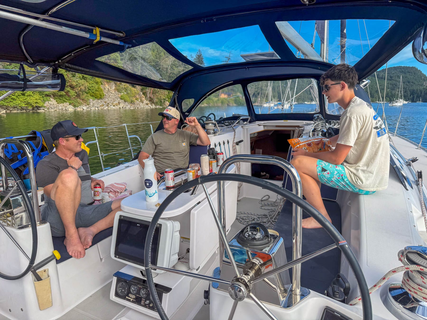 three men relaxing on Simply Dreaming sailboat, surrounded by green hills and other boats under a blue sky