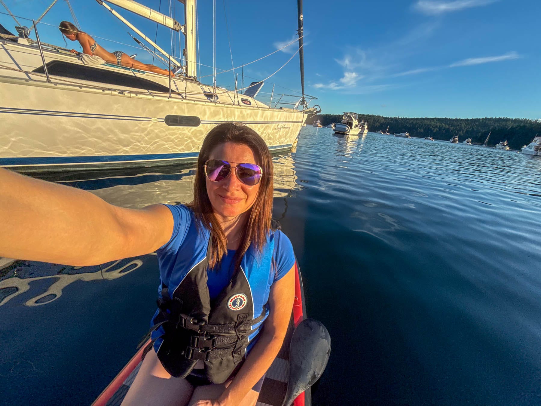 Jami Savage in sunglasses and life vest in kayak, taking a selfie, with a sailboat and other boats in the background