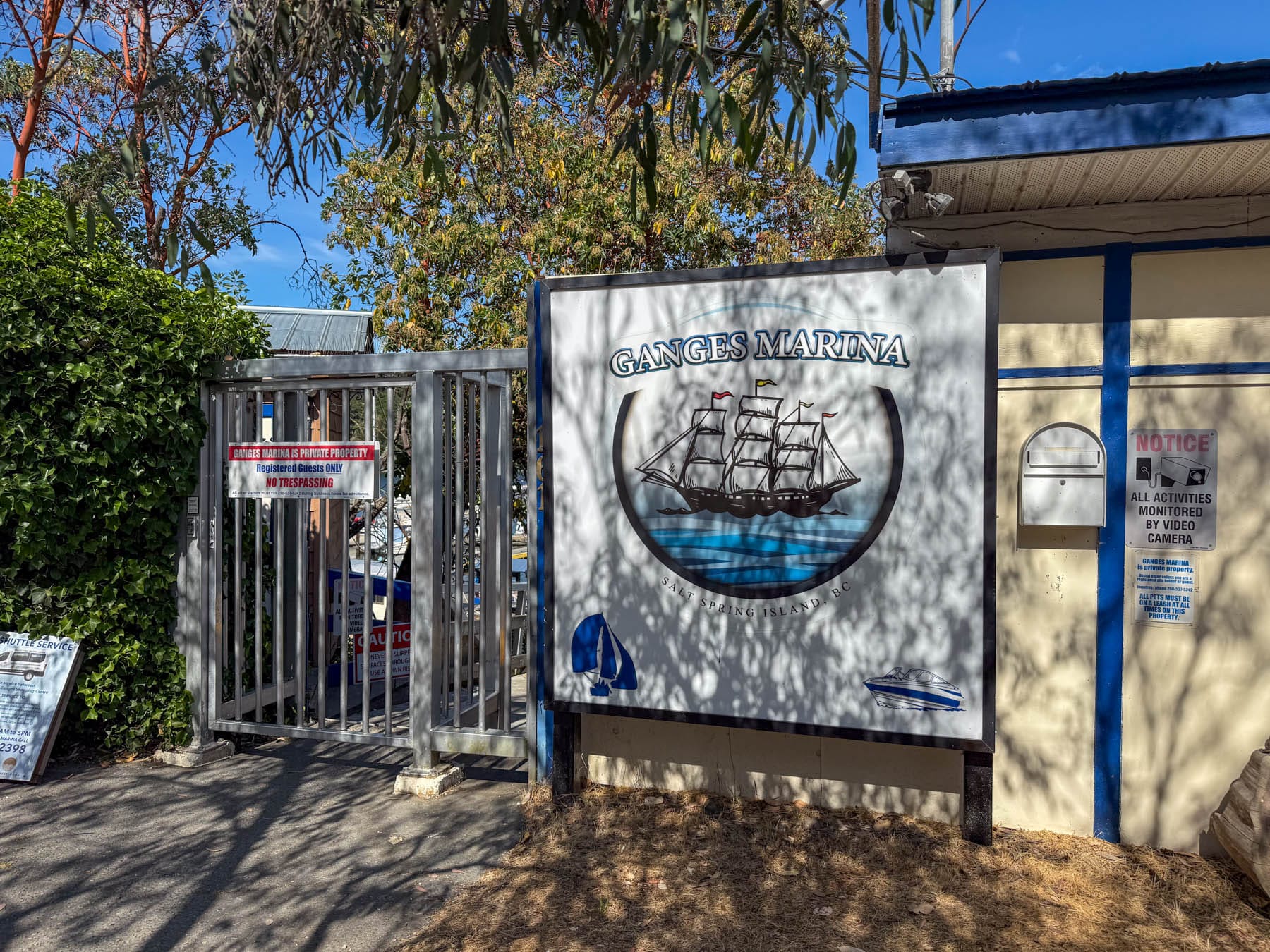 Sign for Ganges Marina with boat logos, private property signs, and a metal gate on a sunny day