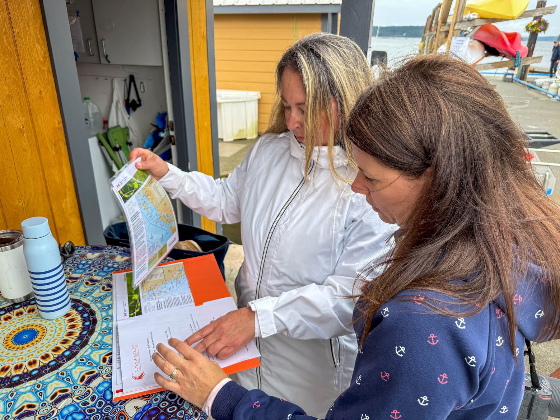 Jami Savage and Tracy Sarich reviewing nautical charts and a guide on a colorful table with a water bottle and mug, next to a dock