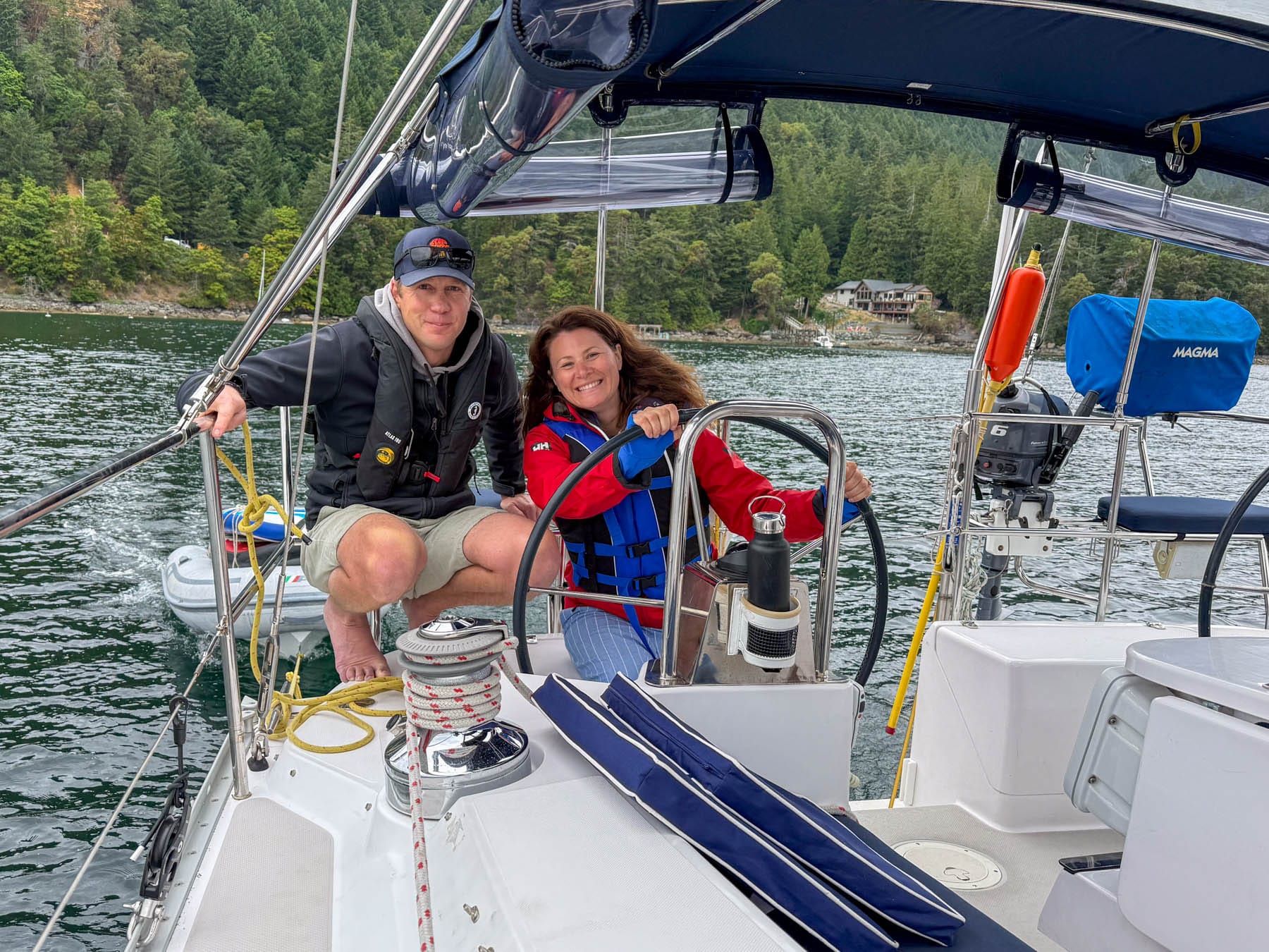 Paul Miller and Jami Savage in life vests on a sailboat, smiling at the camera; forested shoreline and houses in the background