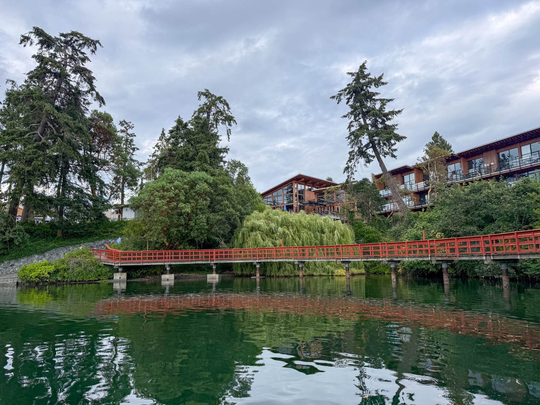 red bridge over dark green water in front of Brentwood Bay Resort and trees on the shore under a cloudy sky