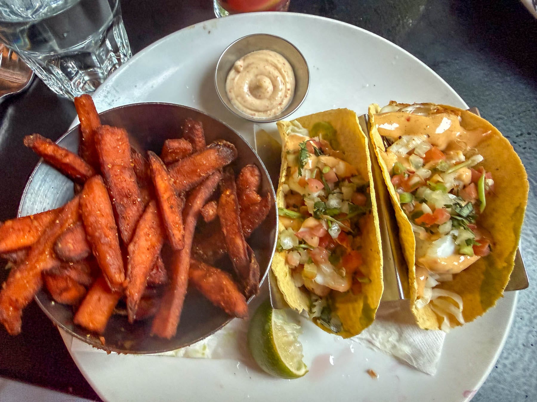 Two tacos and sweet potato fries with dipping sauce and a lime wedge on a white plate