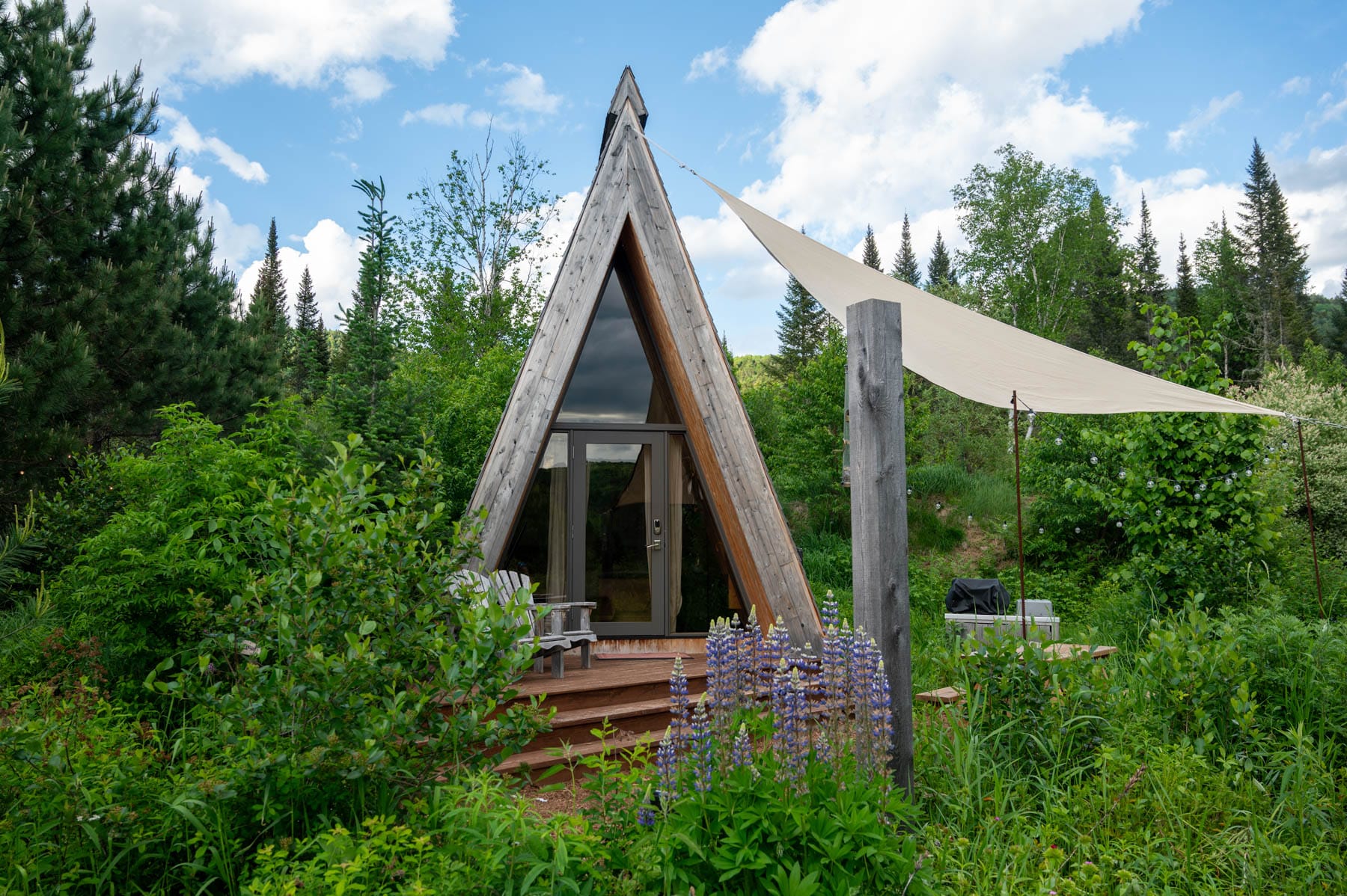 Farouche Tremblant A-frame cabin with a glass door, wooden deck, two Adirondack chairs, surrounded by greenery