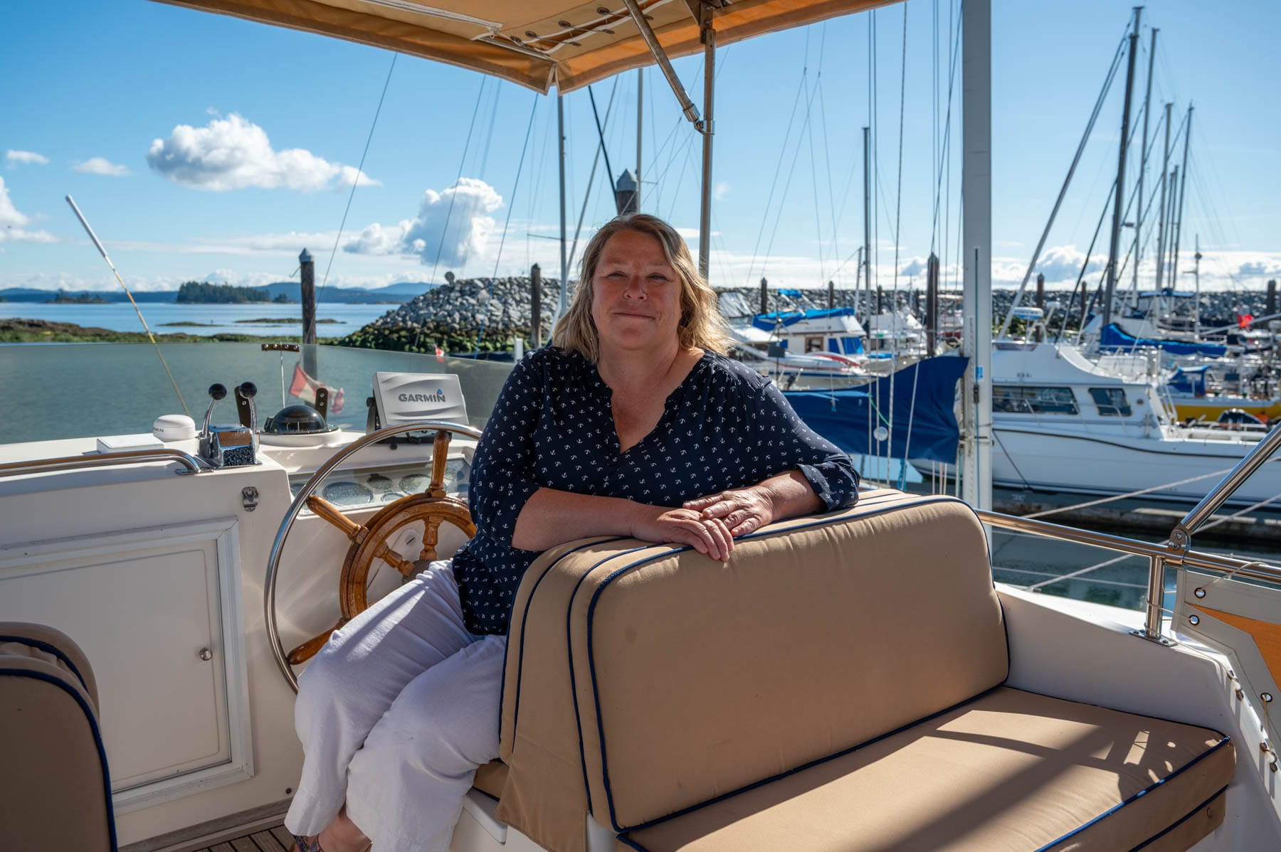 Robin McKeown on boat smiling, with marina and blue sky in background; wooden steering wheel and Garmin GPS visible