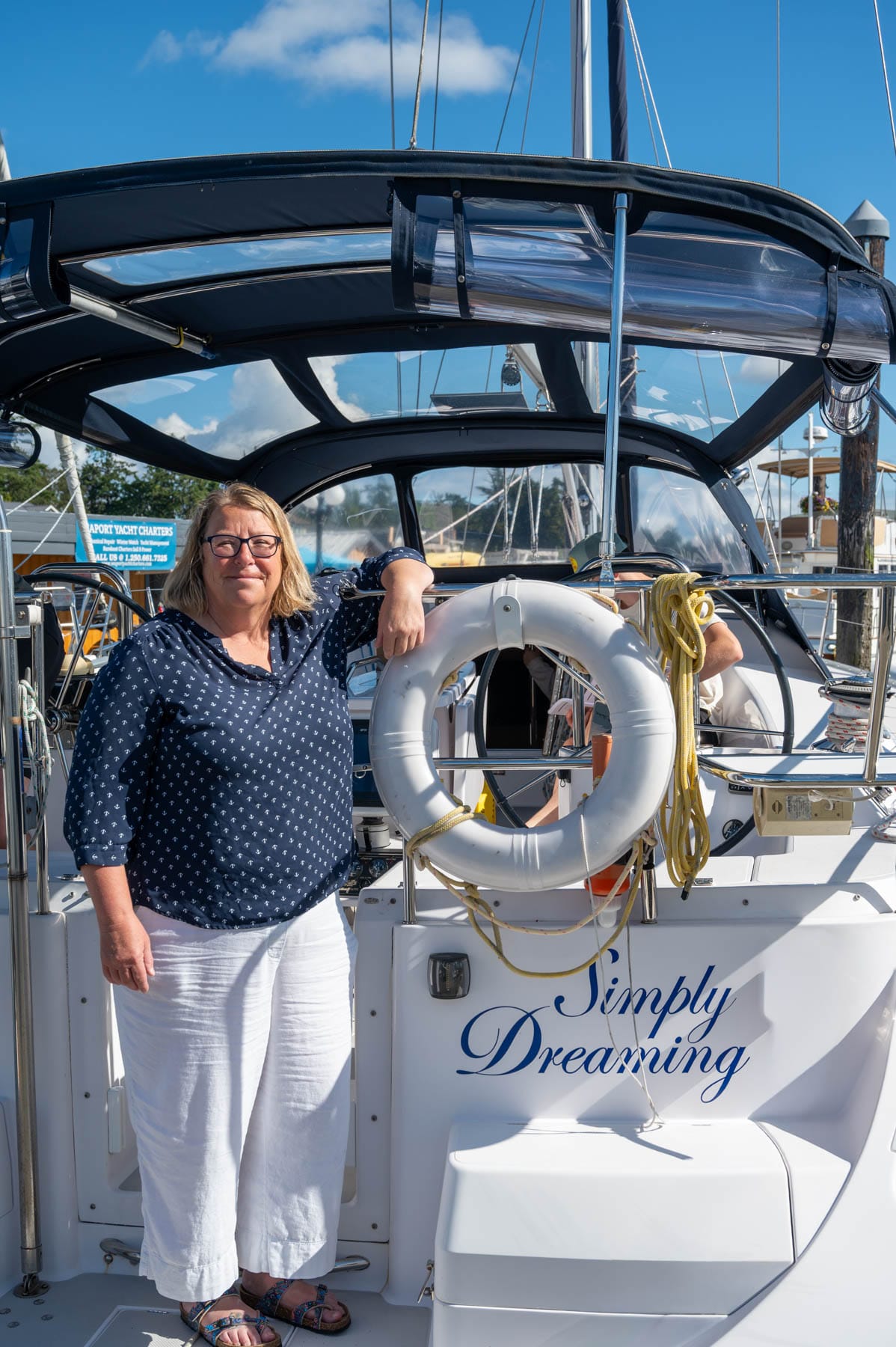 Robin McKeown in blue anchor-patterned shirt and white capris on a white sailboat named "Simply Dreaming" with a life preserver