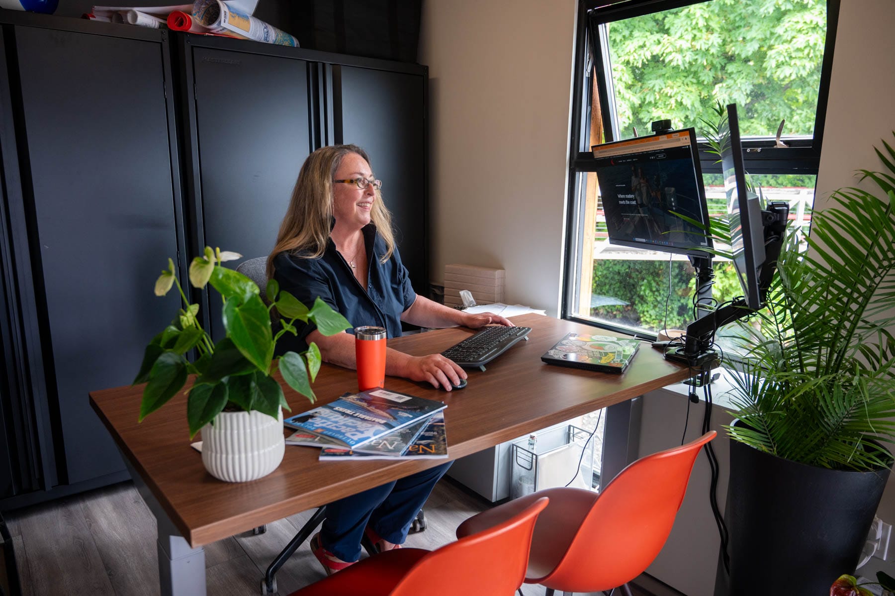 Tracy Sarich into the Voyage Makers Coastal Adventures office, working at a desk with dual monitors, potted plants, and an orange tumbler