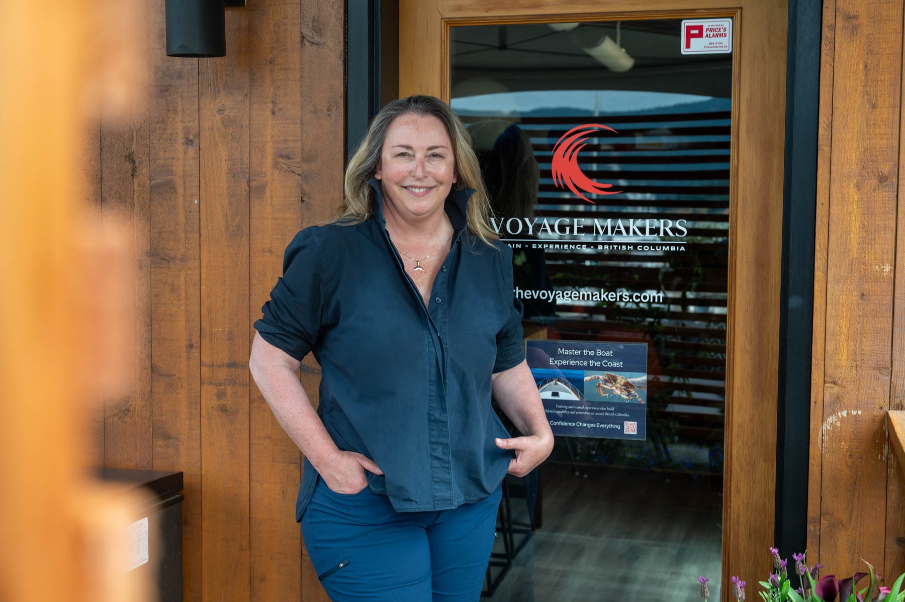 Family Sailing Lessons in Victoria - Tracy Sarich smiling, in a dark blue shirt and blue pants, standing in front of the Voyage Makers Coastal Adventures office