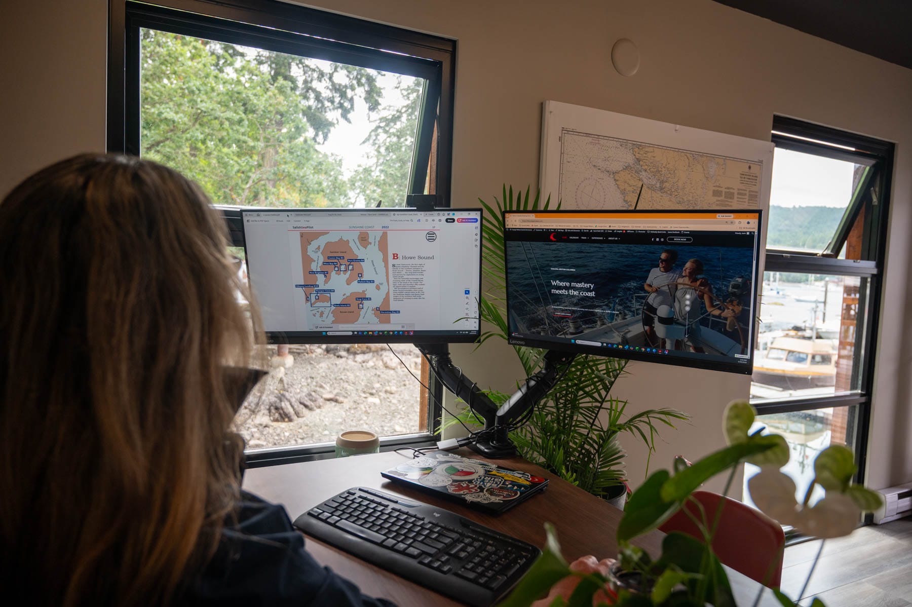 Tracy Sarich viewing marine charts and Voyage Makers website on dual monitors, with a harbor and trees visible through the window