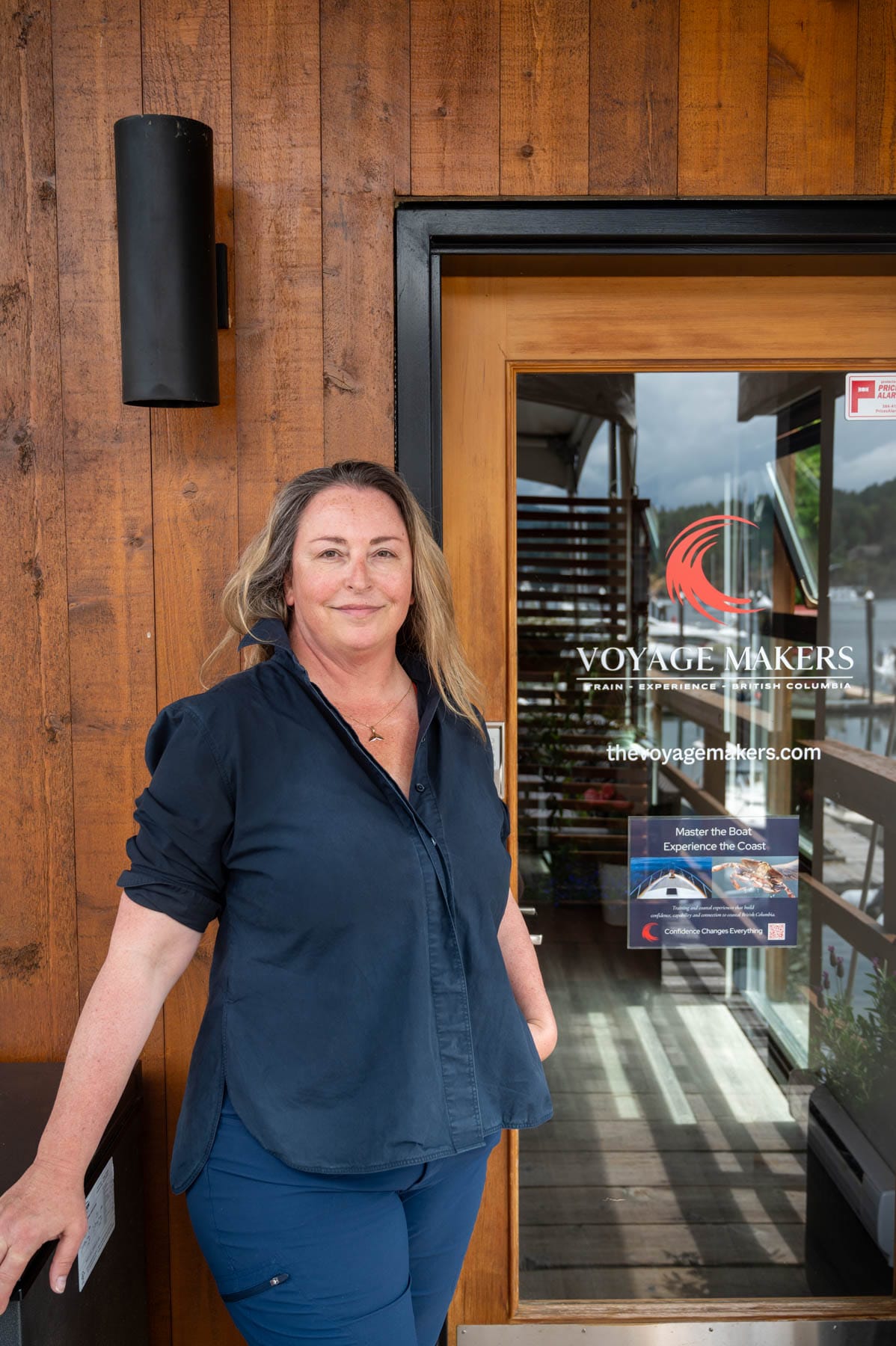 Tracy Sarich smiling, in a dark blue shirt and blue pants, standing in front of the Voyage Makers Coastal Adventures office