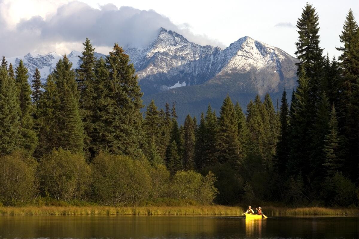 scenic view of two people canoeing on a calm lake with evergreen trees and snow-capped mountains in Bowron Lake Provincial Park, BC, Canada
