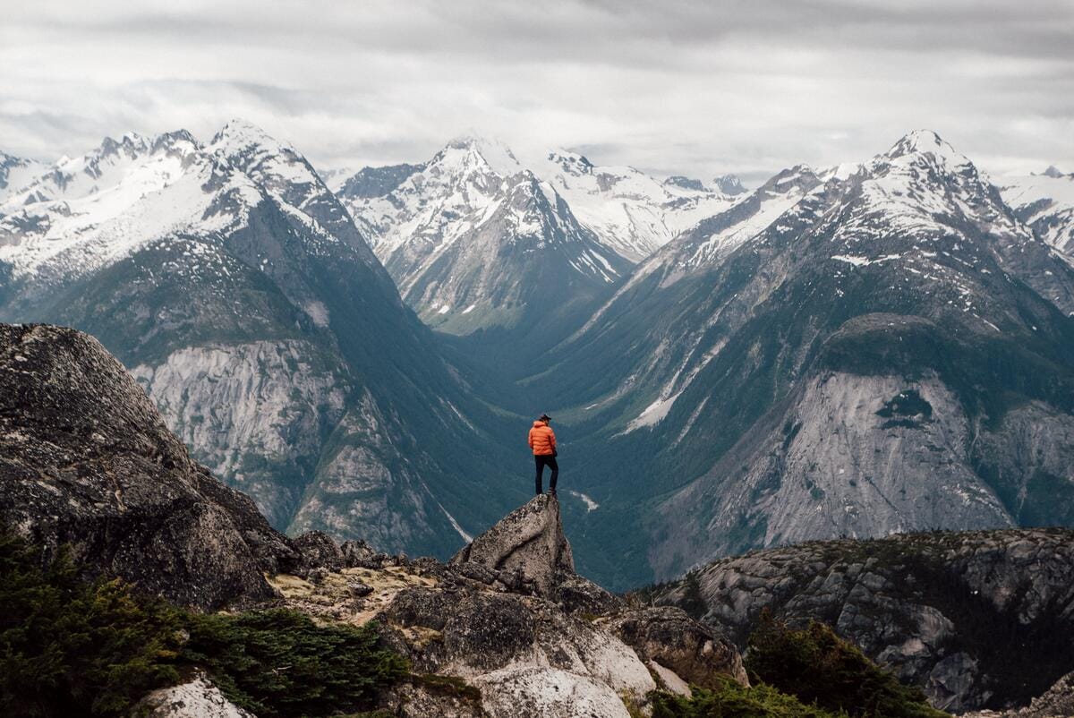 hiker standing on a rocky outcrop overlooking a sweeping, glaciated mountain valley in the Coast Mountains region of British Columbia, Canada