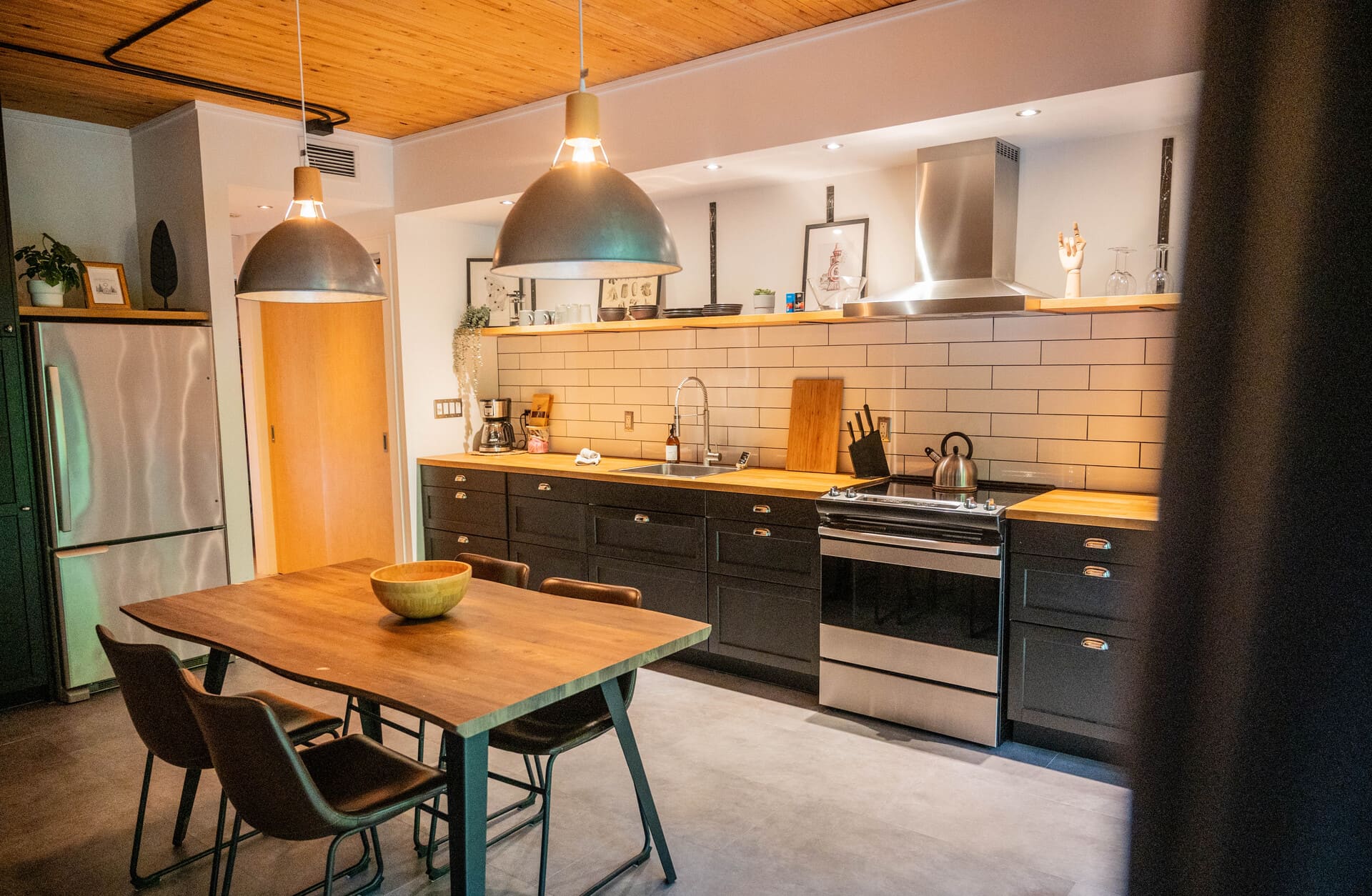 Modern kitchen with black cabinets, wood counters, white subway tiles, and stainless steel appliances. Dining table with three chairs.