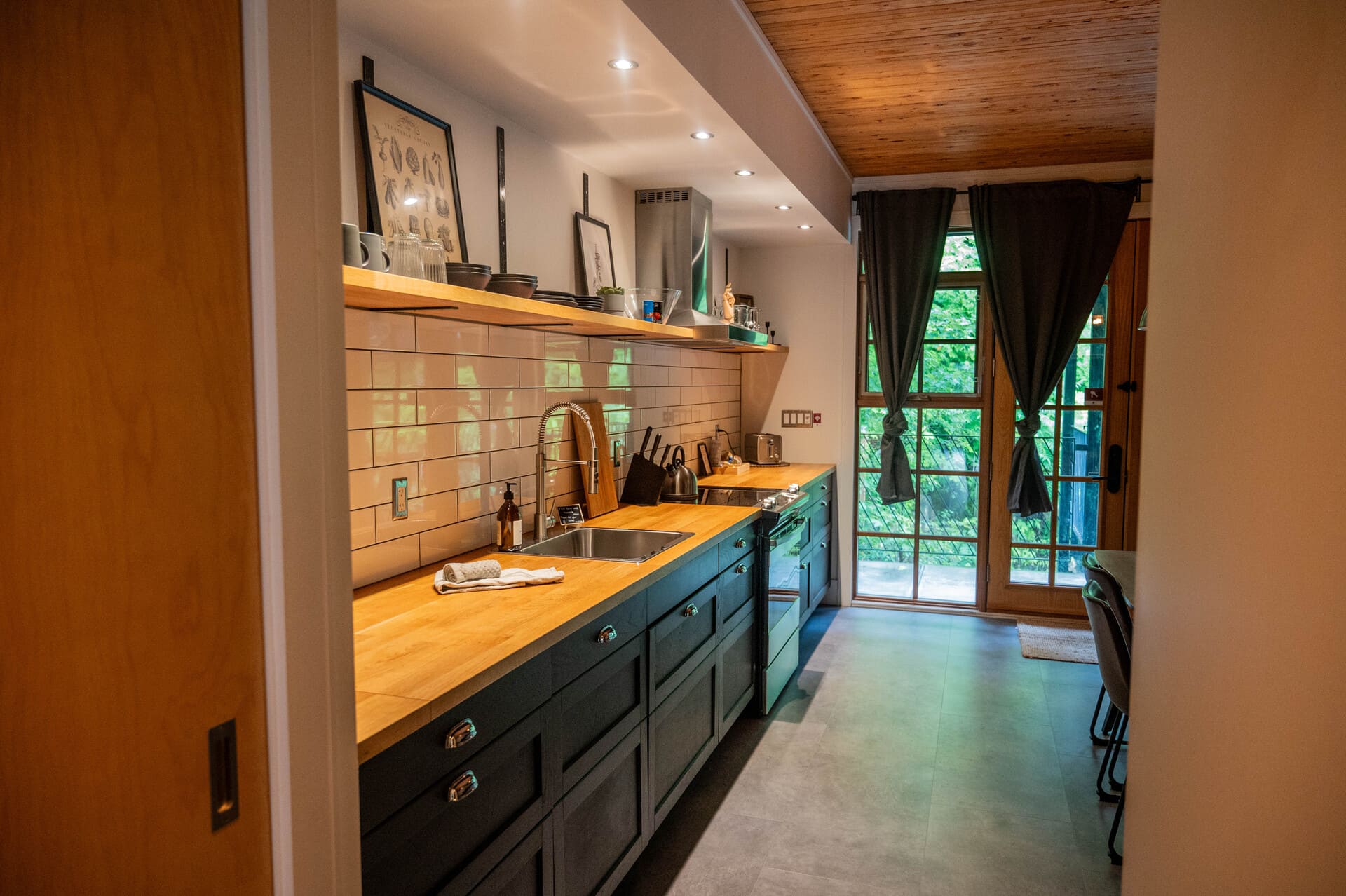 Modern kitchen at "Lofts Du Village" (Chelsea, Quebec) with dark grey cabinets, wood countertops, subway tile, stainless steel appliances, and glass door to patio.