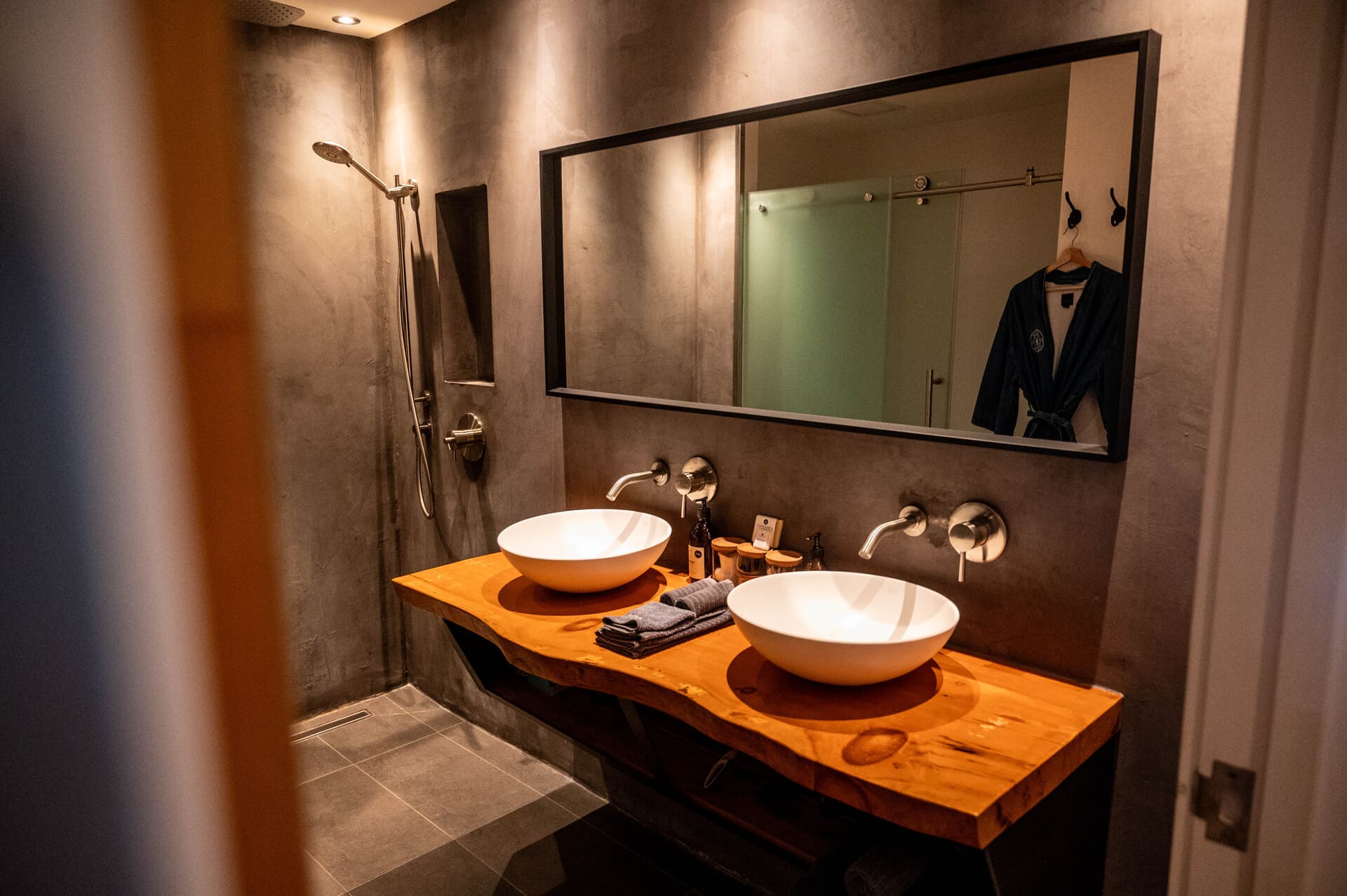 Modern bathroom at "Lofts Du Village" (Chelsea, Quebec)  with double white basin sinks on a live-edge wood counter, gray concrete walls, shower, and large mirror.