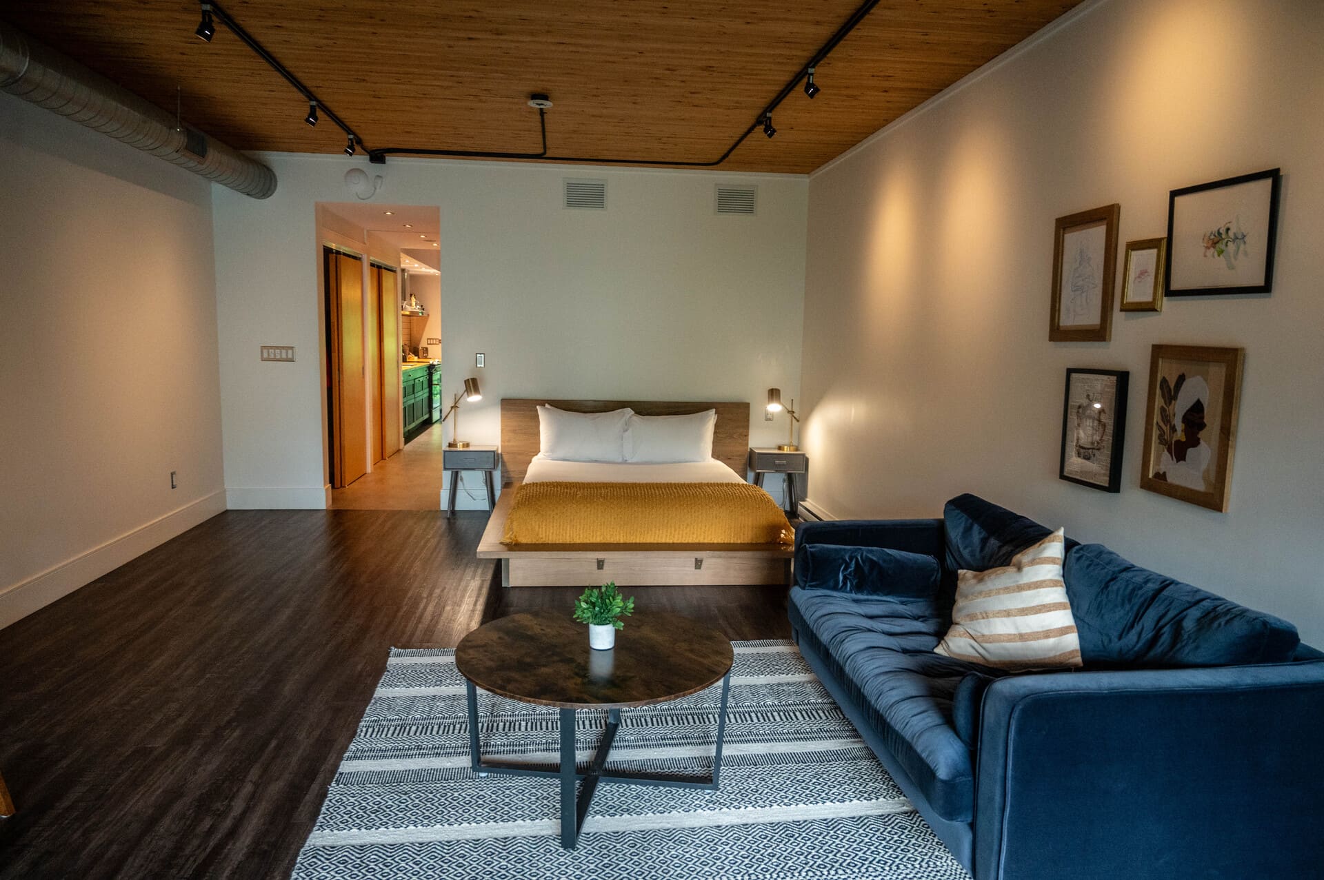 Modern studio apartment with wooden ceiling, dark floors, bed with yellow blanket, navy sofa on geometric rug, and kitchen access.