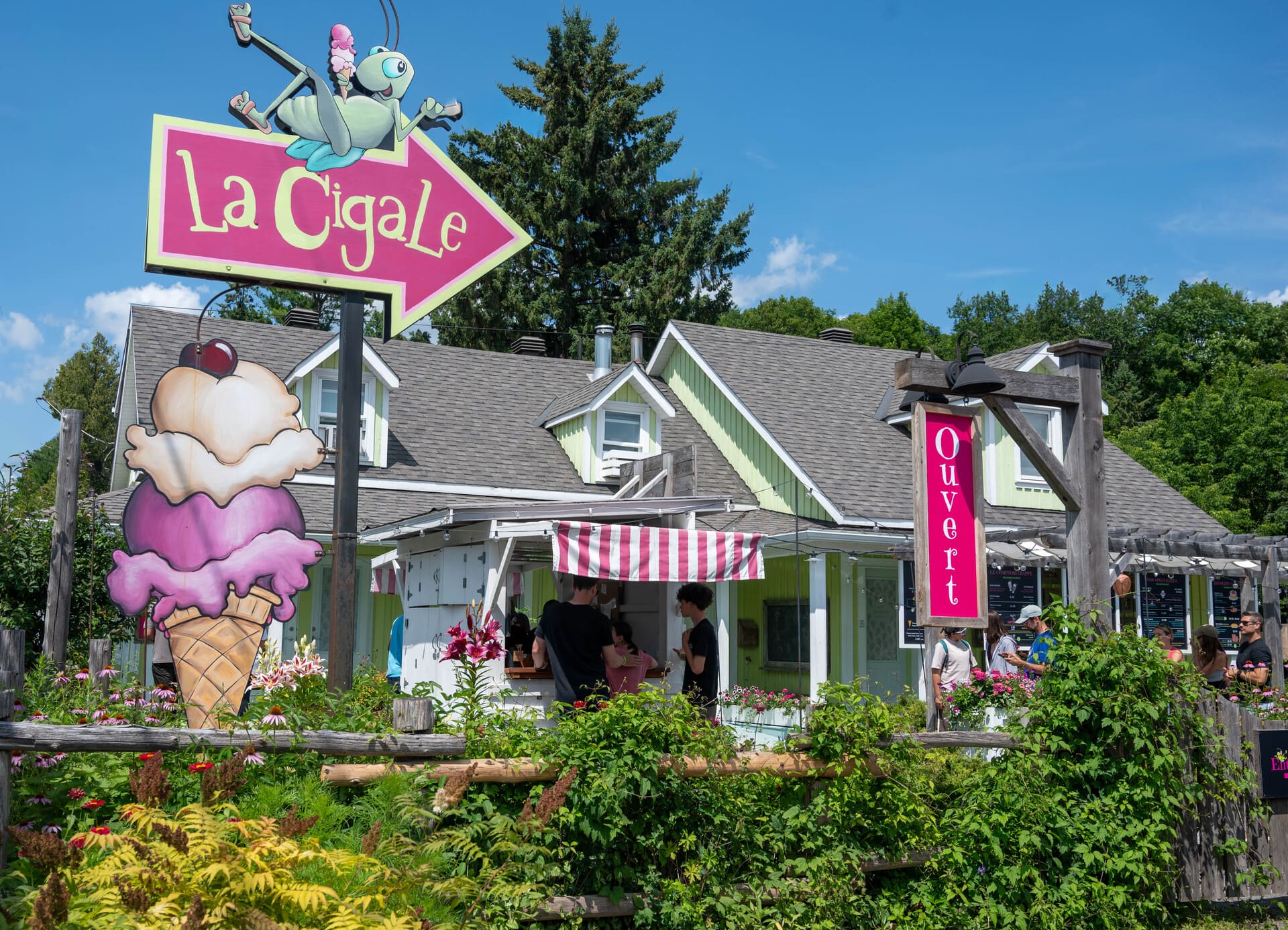 Sign for "La Cigale" ice cream shop with a cartoon grasshopper, giant ice cream cone cutout, and people at an outdoor counter