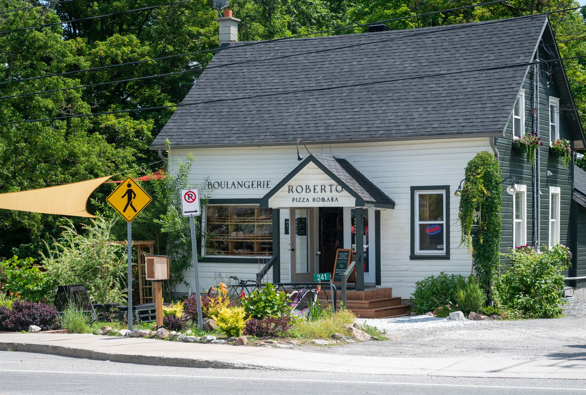 Two-story Boulangerie Roberto building with signage, surrounded by lush greenery, a pedestrian sign, and a paved road.