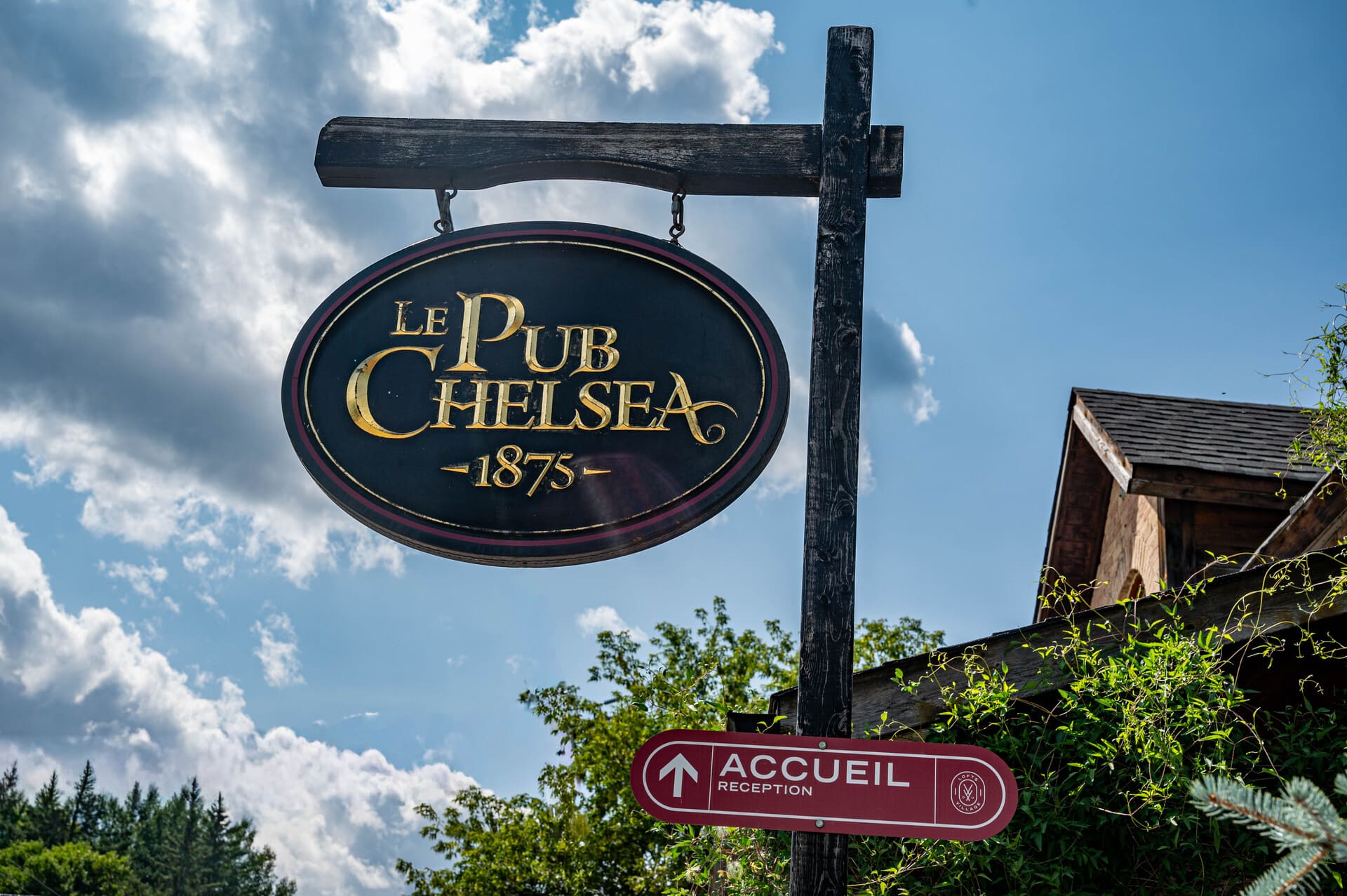 Black oval sign "LE PUB CHELSEA -1875-" and a red "ACCUEIL RECEPTION" sign on a wooden post against a cloudy sky.
