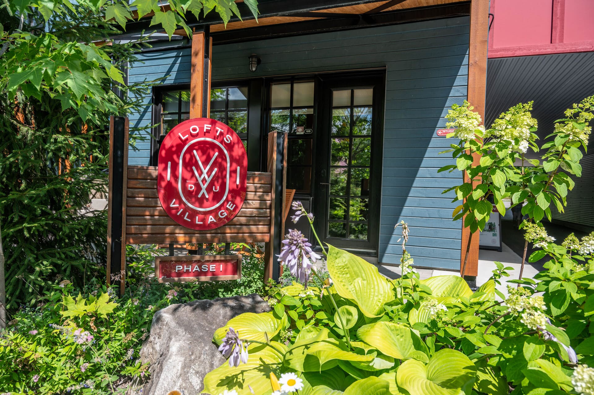 Red "Lofts Du Village" (Outaouais Chelsea, Quebec) sign on a wooden post in front of a blue building with glass doors, surrounded by green plants