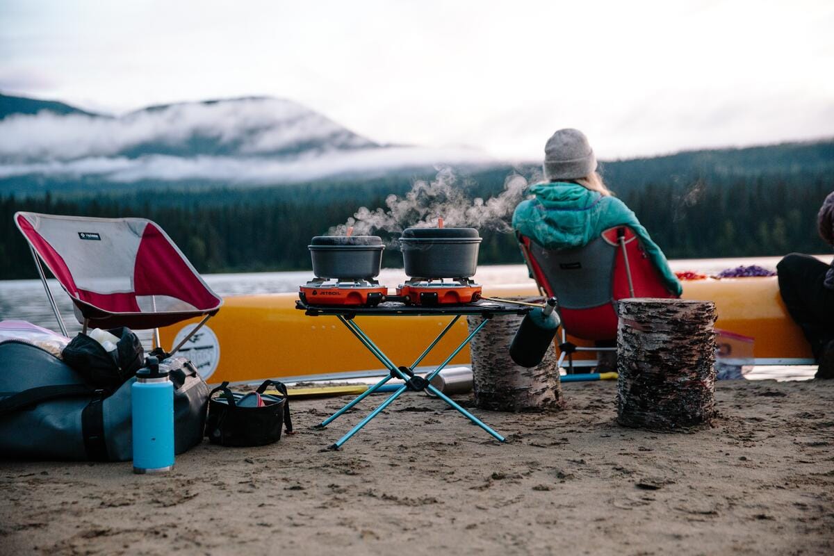 Cariboo Chilcotin Coast - a scenic campsite on a sandy lakeshore with a portable camp stove on a folding table, a person relaxing in a red-and-grey folding chair, and a yellow canoe beached nearby