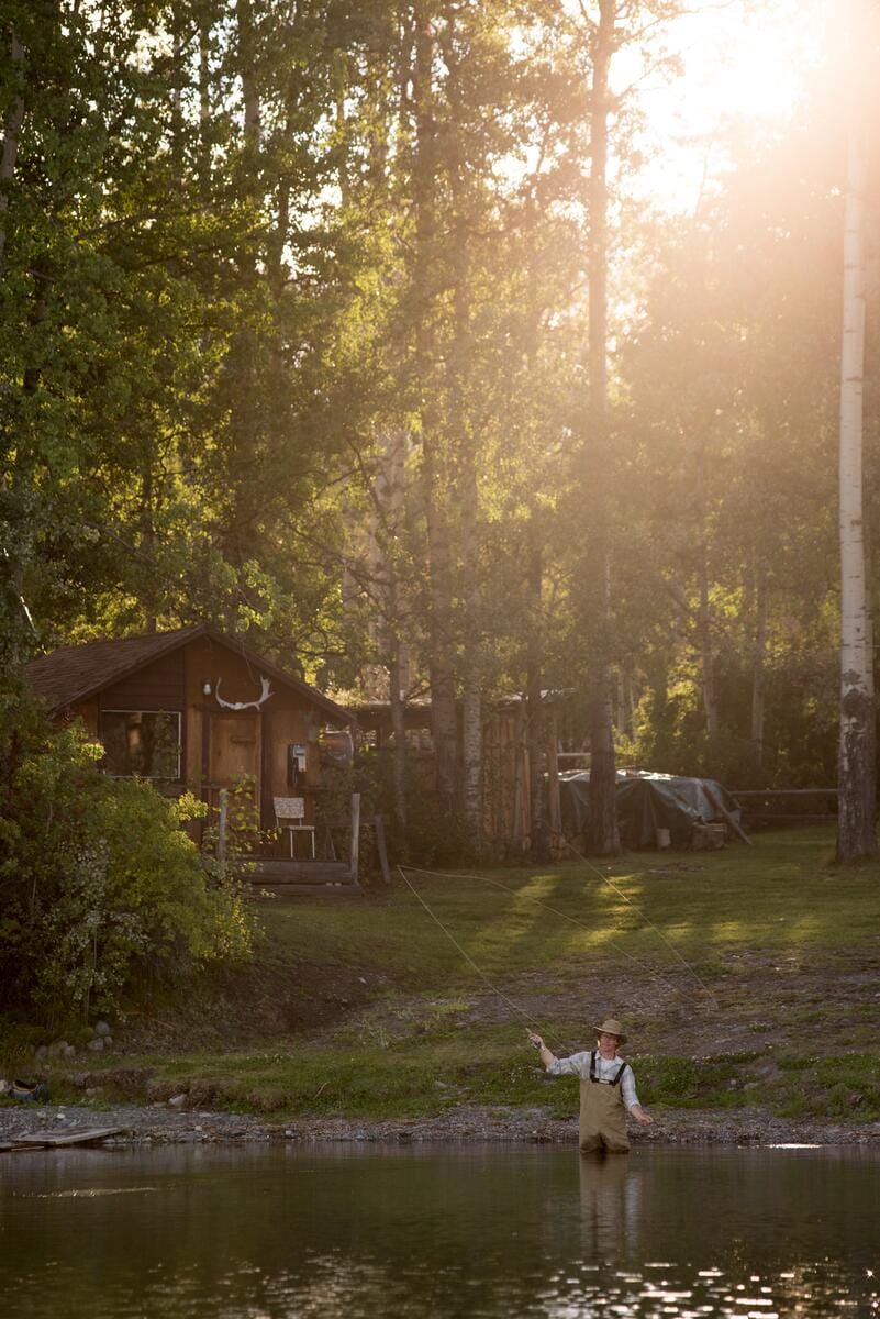 person fly fishing in a shallow river next to a rustic wooden cabin surrounded by tall, sunlit trees