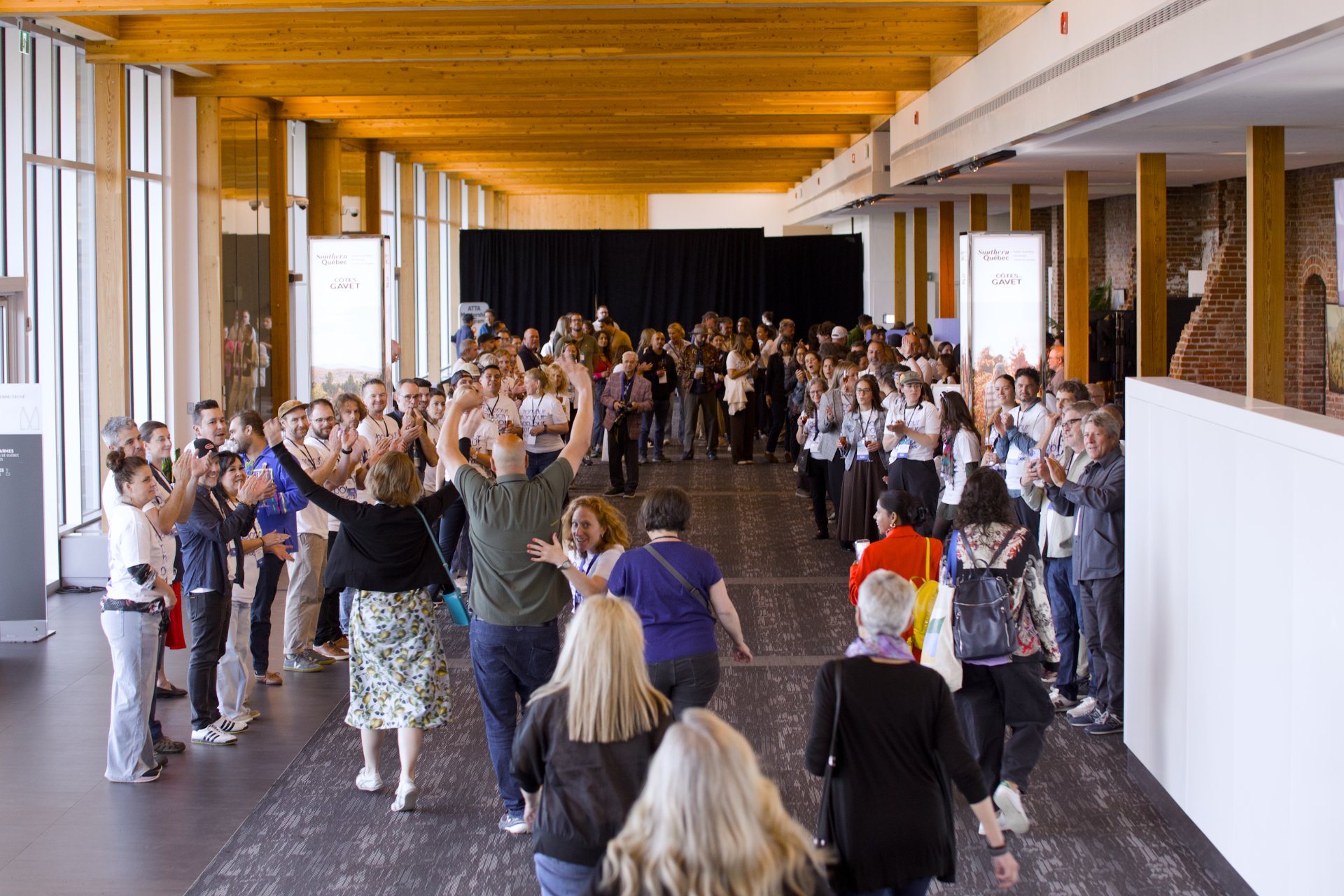 a large group of people walking down a long, light-filled hallway with wooden beam ceilings and large glass windows, resembling a business event space