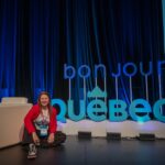 family travel writer Jami Savage sitting on a stage in front of a "bonjour Québec" sign at the Adventure Travel World Summit 2026 (#ATWS2026) in Québec City, Canada