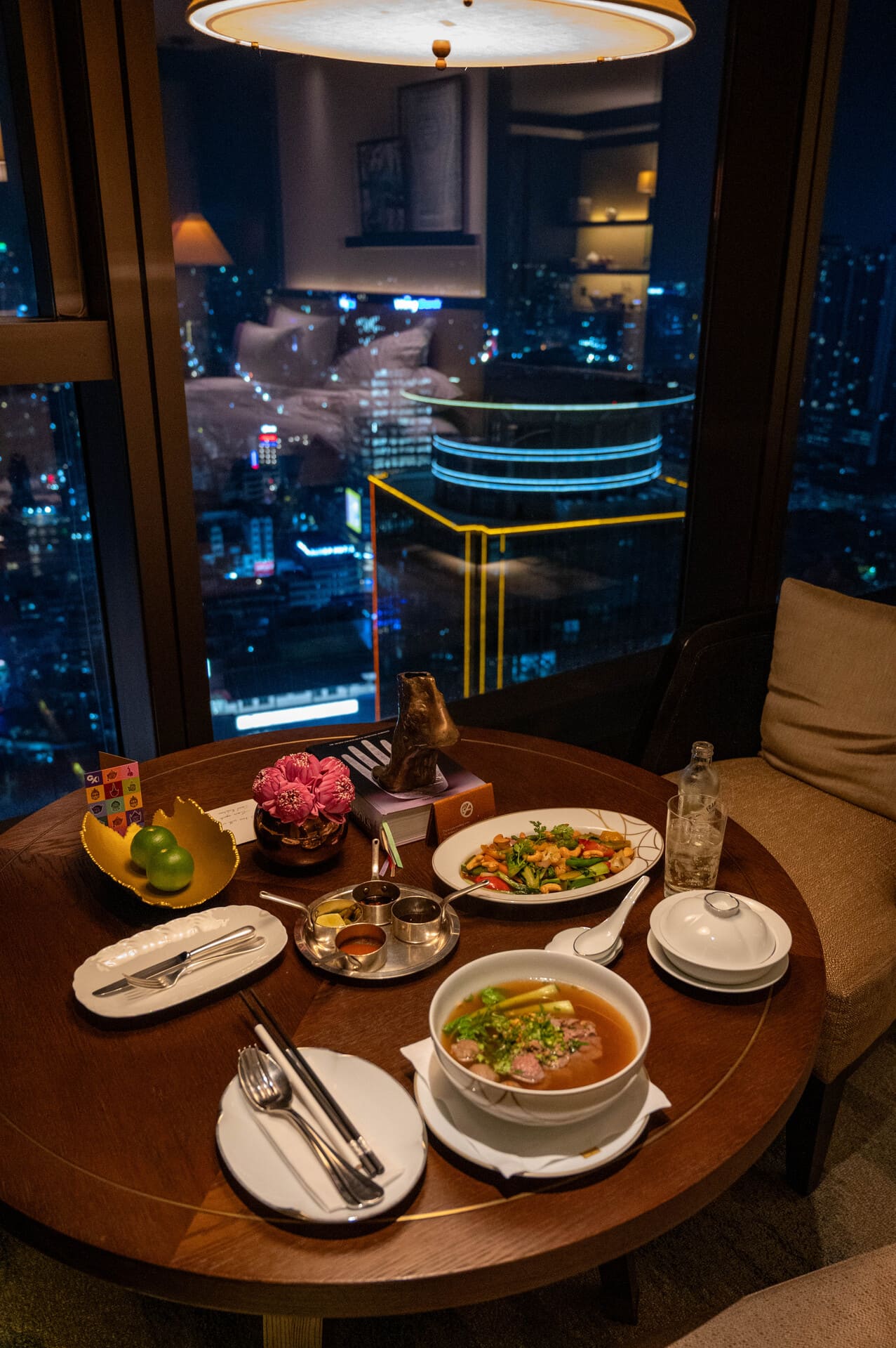 luxury high-rise in-room dining setup on a round wooden table, featuring a bowl of noodle soup/pho, a stir-fried dish, condiments, and floor-to-ceiling windows overlooking a glittering city night skyline