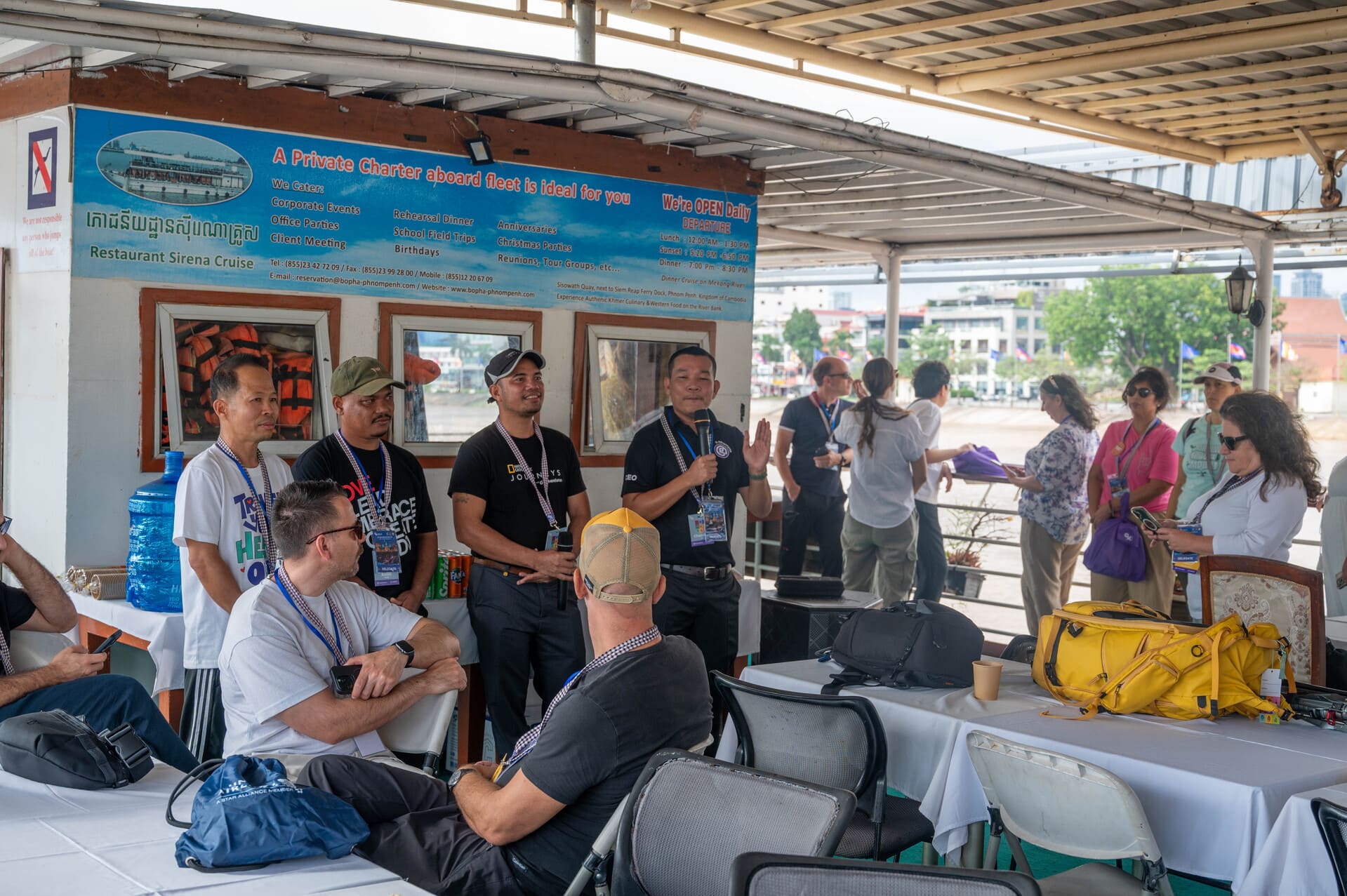 a group of people gathered on a covered river cruise vessel attending a presentation, with banners and signs related to a river cruise (such as Restaurant Sirena Cruise or a Mekong-related program)