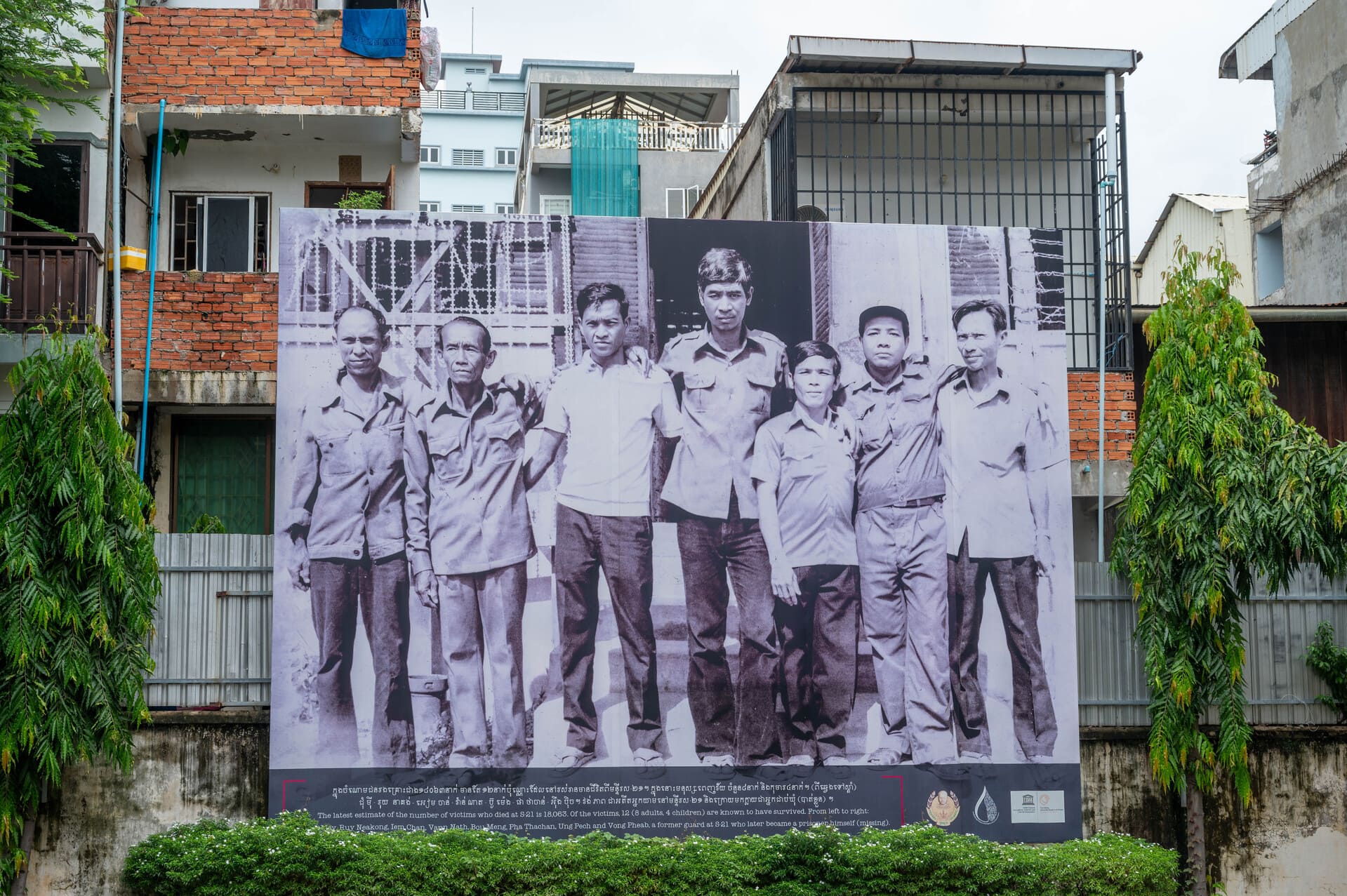 black-and-white memorial banner displays a photograph of survivors from the infamous S-21 (Tuol Sleng) prison camp in Phnom Penh, Cambodia